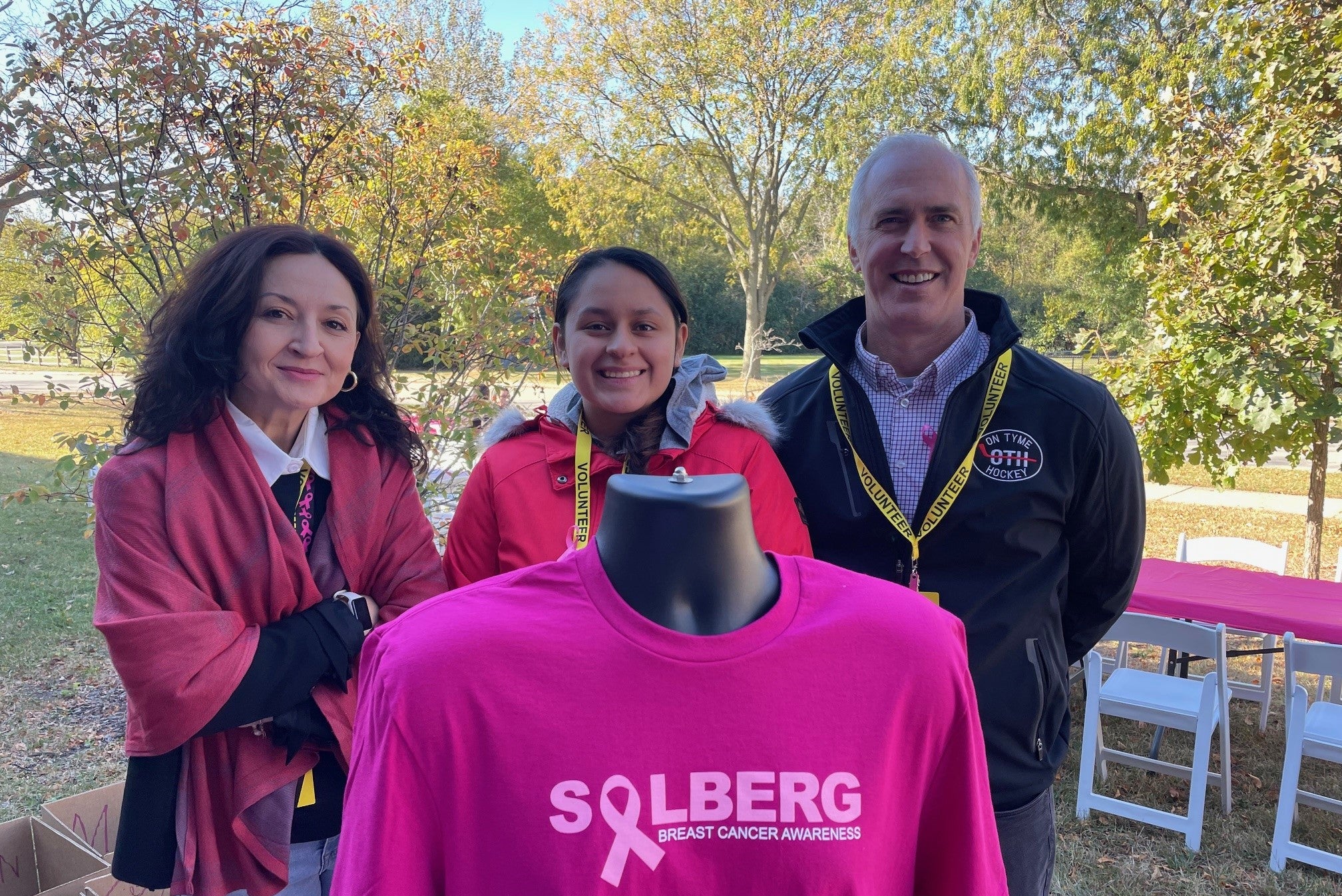 Three people standing outdoors with a pink 'SOLBERG' shirt featuring a breast cancer awareness ribbon.